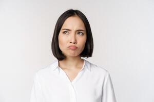 Close up head portrait of young asian woman looking upset and disappointed at left copy space, grimacing and frowning displeased, white background photo