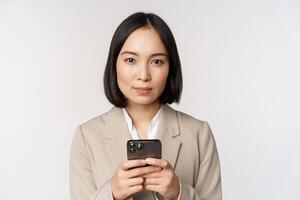 Image of asian businesswoman in suit, holding mobile phone, using smartphone app, smiling at camera, white background photo