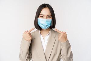 Coronavirus and business people concept. Asian female entrepreneur pointing fingers at medical face mask at workplace, standing over white background photo