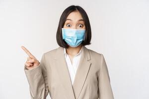 coronavirus y trabajo concepto. retrato de mujer en médico cara mascarilla, señalando dedo izquierda, demostración logo o bandera, anuncio publicitario, blanco antecedentes foto