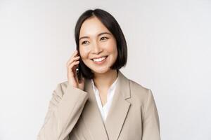 sonriente corporativo mujer en traje, hablando en móvil teléfono, teniendo un negocio llamada en teléfono inteligente, en pie terminado blanco antecedentes foto