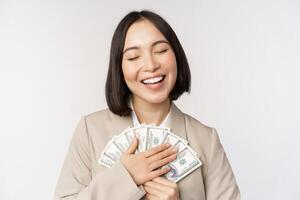 contento asiático mujer de negocios participación dinero en efectivo, abrazando dolares dinero y sonriente, en pie terminado blanco antecedentes en traje foto