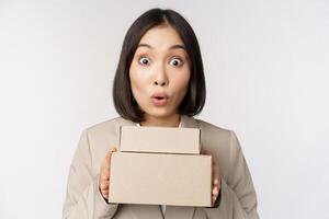 Portrait of asian saleswoman looking surprised, holding boxes, delivery goods, standing amazed in suit against white background photo