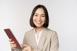 imagen de asiático mujer de negocios, vendedora participación digital tableta y sonriente, trabajando con artilugio, en pie en traje terminado blanco antecedentes foto