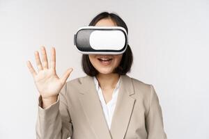 Meeting in vr chat. Asian businesswoman in virtual reality glasses, waving hand and saying hello, greeting someone, standing over white background photo