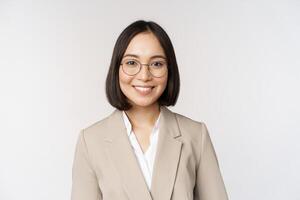 Portrait of young asian saleswoman in glasses, wearing beige suit, smiling and looking confident at camera, white background photo