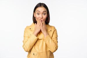 Portrait of excited asian girl looking with interest at camera, amazed reaction at big news or announcement, standing in yellow jacket over white background photo