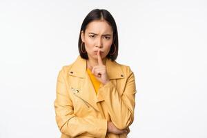 Portrait of angry korean girl shushing, woman frowning and tell to be quiet, showing hush, shh gesture, press finger to lips, taboo sign, standing over white background photo