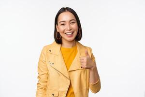 Beautiful korean girl smiling, showing thumbs up, like gesture, recommending store or company, standing satisfied against white background photo