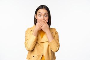 Image of shocked asian girl shuts mouth, close lips and looking speechless, startled face expression, standing over white background photo