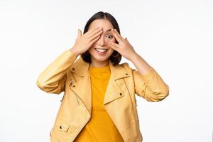 Beautiful, happy asian woman shuts eyes, blindfolded waiting for surprise and smiling, peeking through fingers, anticipating gifts on holiday, standing over white background photo