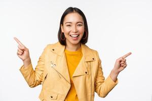 Happy beautiful asian girl pointing fingers left and right, showing banner logo, demonstrating sale, standing in yellow jacket over white background photo