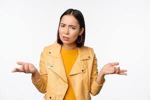 Image of confused young asian woman shrugging shoulders, looking puzzled and clueless at camera, cant understand, standing against white background photo