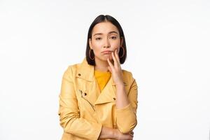 Image of stylish japanese girl looking with unamused, tired and upset face expression, standing in casual clothes over white background photo