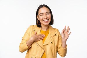 Image of friendly asian girl in stylish yellow coat, raising arm, introduce herself, greeting, waving hand, saying hello, standing over white background photo