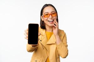 Enthusiastic asian female model, showing smartphone app interface, online store or website on mobile phone screen, standing over white background photo