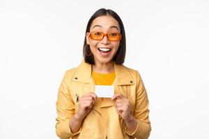 retrato de hermosa moderno asiático niña en Gafas de sol, sonriente feliz, demostración crédito tarjeta, en pie terminado blanco antecedentes foto