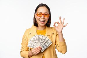 Microcredit and loans concept. Happy stylish korean girl showing okay sign, ok and money, dollars cash, standing in trendy clothes over white background photo