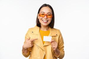 retrato de hermosa moderno asiático niña en Gafas de sol, sonriente feliz, demostración crédito tarjeta, en pie terminado blanco antecedentes foto