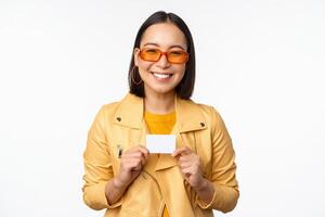 retrato de hermosa moderno asiático niña en Gafas de sol, sonriente feliz, demostración crédito tarjeta, en pie terminado blanco antecedentes foto