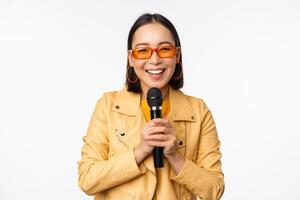 Stylish asian girl in sunglasses, singing songs with microphone, holding mic and dancing at karaoke, posing against white background photo