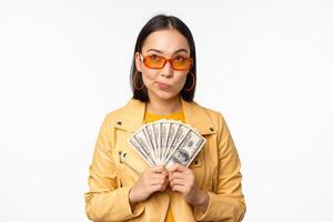 Microcredit and money concept. Stylish asian young woman in sunglasses, laughing happy, holding dollars cash, standing over white background photo