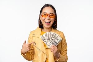 Microcredit and money concept. Stylish asian young woman in sunglasses, laughing happy, holding dollars cash, standing over white background photo