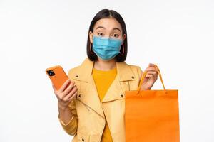 Image of stylish asian girl shopper, holding smartphone and shopping bag without store logo, wearing medical face mask from covid-19, white background photo
