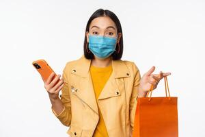 Image of stylish asian girl shopper, holding smartphone and shopping bag without store logo, wearing medical face mask from covid-19, white background photo