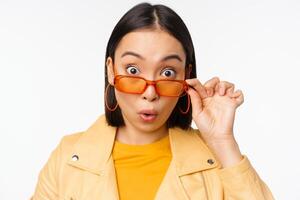 Close up portrait of asian girl looking surprised, wow face, takes off sunglasses and staring impressed at camera, standing over white background photo