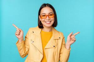 Happy stylish chinese girl in sunglasses, points fingers sideways, invites to check out, see store discount, shows left and right, stands over blue background photo