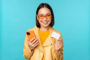en línea compras. elegante joven asiático mujer en Gafas de sol, demostración crédito tarjeta y utilizando teléfono inteligente, pago en Internet, haciendo compra, en pie terminado azul antecedentes foto
