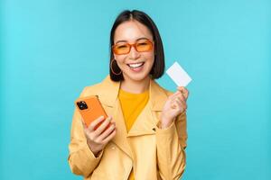 en línea compras. elegante joven asiático mujer en Gafas de sol, demostración crédito tarjeta y utilizando teléfono inteligente, pago en Internet, haciendo compra, en pie terminado azul antecedentes foto