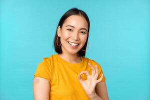 Vaccine campaign from covid-19. Happy healthy asian girl showing okay sign and smiling, bandaid on shoulder from coronavirus vaccination, blue background photo