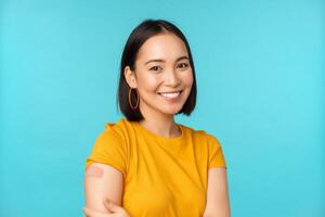 Vaccine campaign from covid-19. Young beautiful, healthy asian woman showing shoulder with bandaid, concept of vaccination, standing over blue background photo