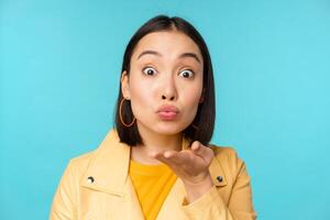 Close up portrait of funny asian girl sending air kiss, blowing at camera with popped eyes, standing over blue background photo