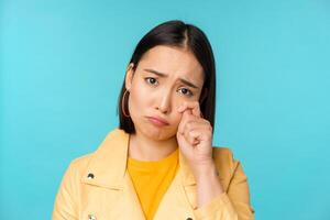 Close up of sad asian girl crying, wiping tears and sulking, looking upset and gloomy, posing against blue background photo