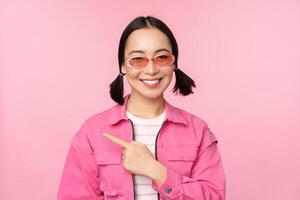 Portrait of smiling asian girl in stylish outfit, sunglasses, pointing finger left, showing advertisement, banner, standing over pink background photo