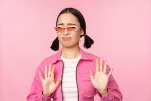 No thanks. Disgusted asian girl in sunglasses, rejecting smth, shaking hands to refuse, decline, standing over pink background photo