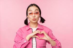 Lovely korean female model in trendy sunglasses, shows heart, care sign, I love you gesture, stands over pink background photo