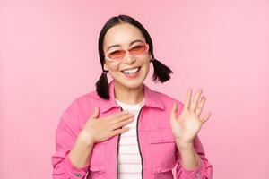 Portrait of friendly asian teen girl in sunglasses saying hello, waves her hand and smiles, greets you, hi gesture, stands over pink background photo