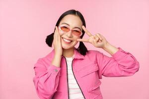 retrato de kawaii asiático niña en Gafas de sol, demostración paz, signo v cerca ojo y sonrisas linda a cámara, posando en de moda ropa en contra rosado antecedentes foto