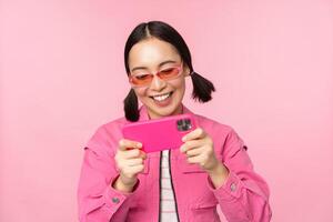 Portrait of happy asian girl playing on smartphone, watching videos on mobile phone app, standing over pink background photo