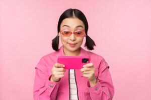 Portrait of happy asian girl playing on smartphone, watching videos on mobile phone app, standing over pink background photo