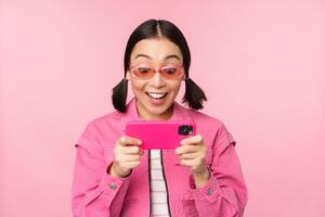 Portrait of happy asian girl playing on smartphone, watching videos on mobile phone app, standing over pink background photo