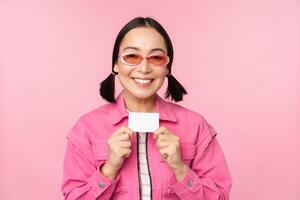 Portrait of stylish, modern asian girl, shows discount, credit card and look pleased, paying contactless, concept of shopping, standing over pink background photo