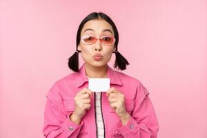 Portrait of stylish, modern asian girl, shows discount, credit card and look pleased, paying contactless, concept of shopping, standing over pink background photo