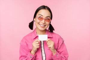 Portrait of stylish, modern asian girl, shows discount, credit card and look pleased, paying contactless, concept of shopping, standing over pink background photo