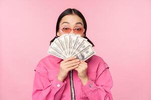 Beautiful korean woman in sunglasses, showing dollars, money cash, smiling pleased, concept of fast loans, microcredit and payment, standing over pink background photo