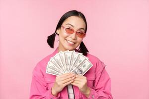 Beautiful korean woman in sunglasses, showing dollars, money cash, smiling pleased, concept of fast loans, microcredit and payment, standing over pink background photo